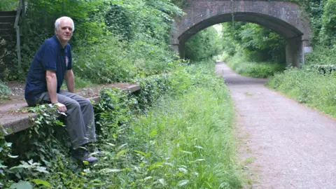 Tom Carswell Tom is sat on a train platform with his legs hanging over the edge into weeds. The track has been removed from the line and is instead a pathway. A tunnel runs over the track.