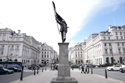 PA Media A grey statue of a man wearing a suit, walking forward off a plinth, carrying a flag that covers his face