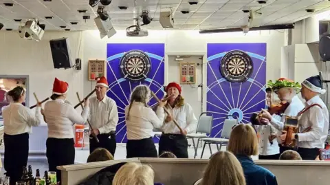 A group of five Morris dancers in white shirts, black trousers and red Christmas hats dancing with sticks in front of an audience. Three other members of the band are playing music. There are dartboards on the wall behind them. 