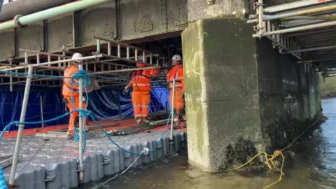 Network Rail Three workers in orange high visibility overalls and white hard hats can be seen on a pontoon underneath the bridge. 