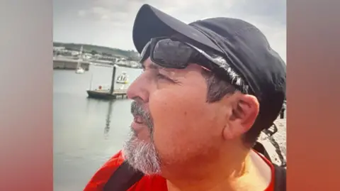 Police handout image of Claudio Aquilino. He is in his 50s and has a greying goatee beard. He is wearing a black baseball cap, dark sunglasses, a red shirt and a black rucksack. He is standing at a harbour.