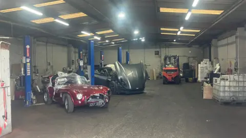 Inside a garage housing cars in the scrapyard. There are two classic style cars and a forklift truck. The warehouse otherwise appears fairly empty with fluorescent lights.