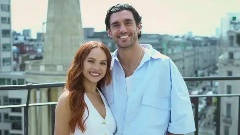 BBC Alexis Warr, a woman in a white top with long, straight red hair, and Julian Caillon, a man in a blue shirt with dark hair slicked back, stand smiling on a balcony with a view of a street with cars and buildings