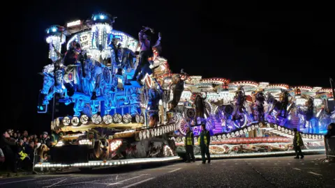 Neil@nodpics A long, brightly-lit carnival cart with people hanging off it all the way down its length. It is photographed from the side.