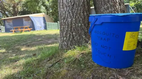 An image of a blue bucket with 'Mosquito trap do not touch written on it' and a yellow food label on the side, sits in front of a light brown tree trunk. In the background, a blue and tawny tent with a picnic table in front. 