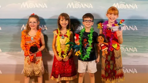 James Gillham/Disney Four children wearing Moana outfits and smiling while standing in front of a Moana backdrop. They also have colourful floral necklaces on.