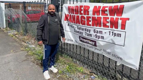 Kevin Shoesmith/BBC Thinesh Rajavatnam next to his banner that states 'under new management'