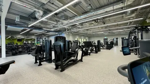 Magna Vitae A gym featuring an array of exercise equipment, including cardio and resistance machinescise machines in a large, well-lit room, with mirrors lining the walls.