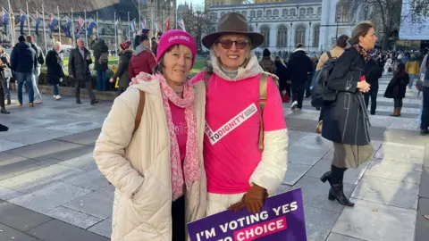 Two women, one wearing a pink t shirt and brown hat, and another in a pink hoody and white jacket. Both are wearing clothes with slogans in support of the assisted dying bill 