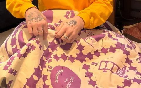 Stewart Francis Easton The hands of Stewart Francis Easton can be seen sewing a large quilt. Purple images are being sewn onto a cloth background. The cuffs of his yellow top can be seen and he has tattoos on his hands, one of a bear the other says Peace. He is also wearing rings. 