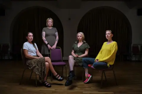 BBC Scotland Four women, three seated and one standing behind a chair. They are in a dark room, looking directly at the camera. 