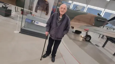 Supplied John standing in front of the aircraft display. He is wearing a grey jacket and holding his walking stick. The Spitfire is in the background, along with a display exhibiting a flying jacket.