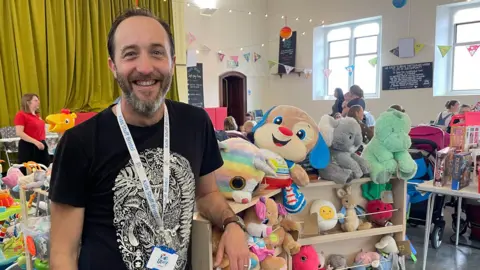 BBC Kyle Henderson-Begg wears a black T-shirt with a white leaf design on it. He stands in front of a rack of stuffed toys. Women holding children are visible in the background. 