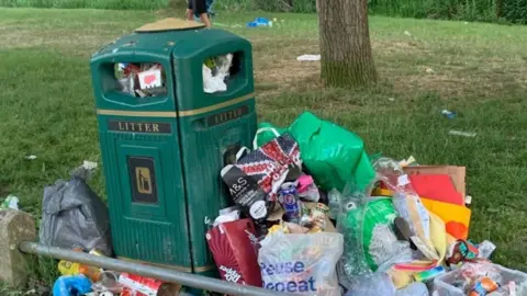 A close up photo of a green bin with the word litter on the site, which has several plastic bags bursting with rubbish propped against it. It is in a grassy location and someone's legs are seen walking behind.