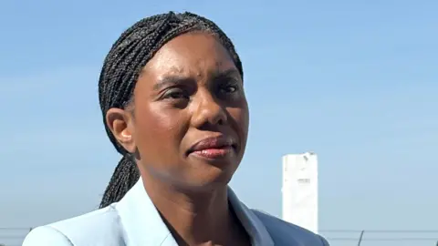 A close-up photograph shows Kemi Badenoch outdoors, looking slightly past the camera, with braided hair pulled back, wearing a light blue jacket. A pale blue sky and a tall white structure are visible in the background.