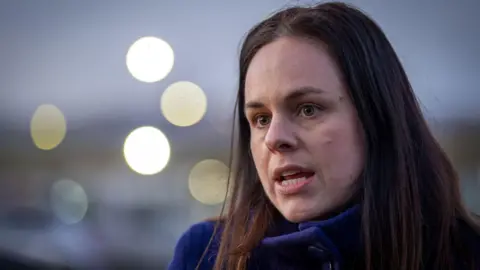 PA Media Kate Forbes has dark hair to her shoulders and is wearing a blue jacket. She is standing outdoors and there are blurred lights in the background.