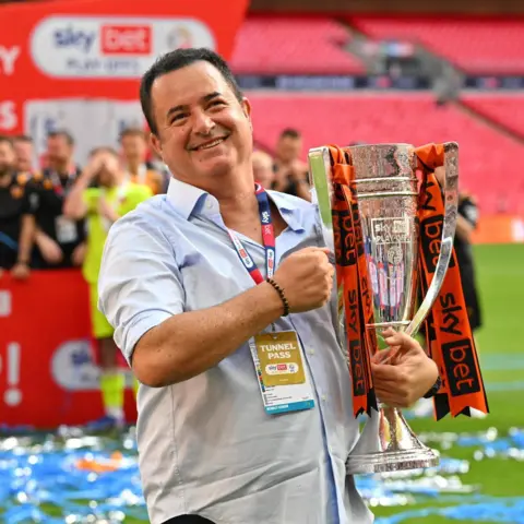Acun Medya A man stands on a football pitch holding a large trophy with orange and black ribbons attached. Advertising boards and people are visible in the background.