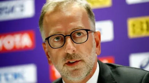 Primer plano de Ed Warner, con traje y gafas, hablando en una conferencia de prensa con un cartel publicitario morado desenfocado detrás de él.