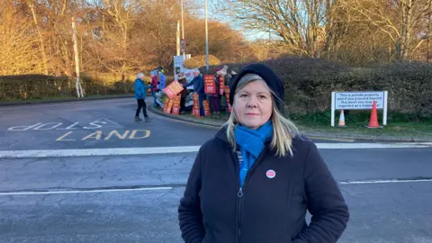 Luke Deal/BBC Alison Downes standing outside the Sizewell site. She is in a dark coloured fleece, blue scarf and a dark coloured hat. Behind her are protestors holding  placards citing 'stop sizewell C'. 