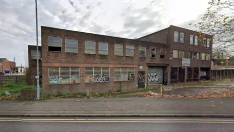 A Google street view image showing a derelict brick-built factory. The windows are dirty and there is graffiti on parts of the building. Some of the windows are missing and a lamppost stands outside it.