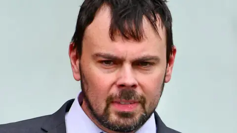 A man wearing a grey suit, purple shirt and tie with white diamonds on it. His top button is undone. He has black hair and hazel eyes and a dark goatie style beard. The background behind him is blue and he is looking down. 