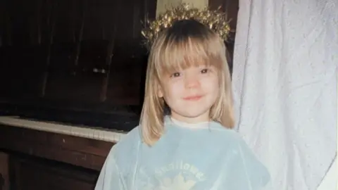 Amy Anderson Amy as a young child wearing a light blue top and a tinsel crown