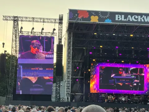 A shot from the crowd of the Stevie Wonder concert in Cardiff. His face, wearing a black hat and sunglasses are on a screen on the left of the shot and in the middle of the stage. The letters Blackw appear at the top of the stage