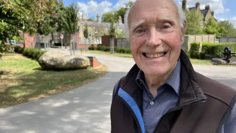 BBC Jack Hayward smiles in a park with the railway village behind him and the DMJ tower in the distance