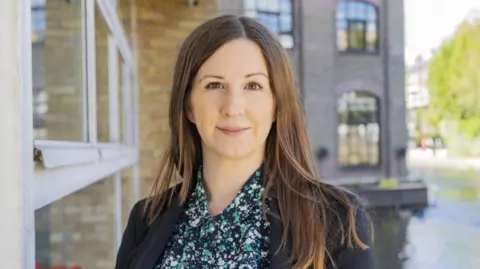 Naomi Clayton The head and shoulders of a woman with shoulder-length brown hair, wearing a dark blazer jacket over a white, grey and navy blouse, standing outside next to a building smiling at the camera.