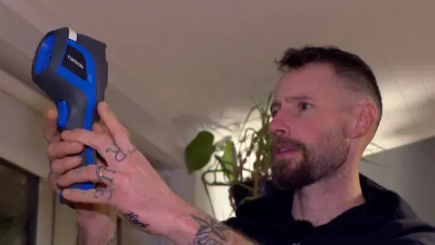 Orion Spalton has short dark hair and a beard and is wearing a black fleece. He's holding a blue and grey thermal imaging camera in his hands looking at the image on the back of it and what it reveals about cold spots on the wall he is pointing it at.