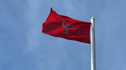 A red Manx Flag flying in the breeze with a blue sky behind it.