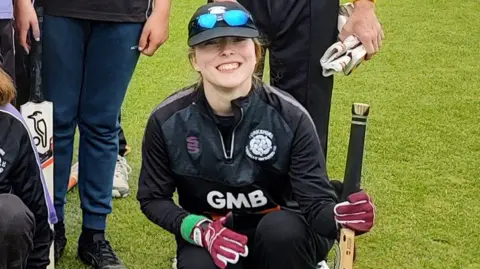 Nick Lowe Jess Lowe a young woman wearing a cricket kit and gloves while carrying a bat. She is kneeling on grass in front of other players. 