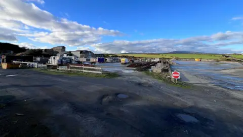 IOM GOVERNMENT An industrial site with a large warehouse with vehicles and containers in front of it on a clear day. There are puddles on the grounds and a No Entry sign at the front of the site, and a blue sky with white clouds above.