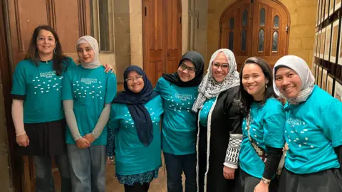 Shariqua Ahmed/BBC Women volunteers, all wearing a teal-coloured Ramadan Tent Project T-shirt, smiling for camera.
