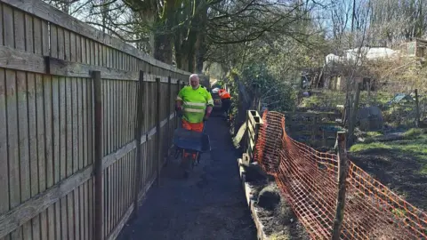 A man in a high-vis jacket moves a wheelbarrow down a footpath. On the left is a fence on the right are allotments 