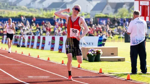 Vannin Photos Corrin Leeming grits his teeth as he crosses a finish line of an orange track. He punches the air with one hand. He wears a red vest and large sunglasses. You can see people watching on a grassy bank.