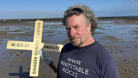Whitstable Oyster Fishery Company Farm Manager Renny Hall stands in the sand about a quarter of a mile off the beach at low tide with a marker buoy behind him which reads "Danger Oyster Trestles"