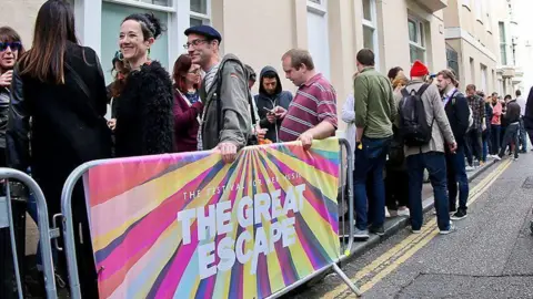 A queue of people outside a building. A metal railing with "The Great Escape" written on it separates the crowd from the road.