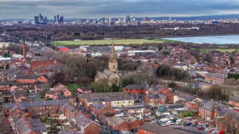 Getty Images Aerial view showing the area with a church surrounded by redbrick terraced houses and field and reservoir behind. Manchester city centre can be seen in the backdrop with its skyscrapers.