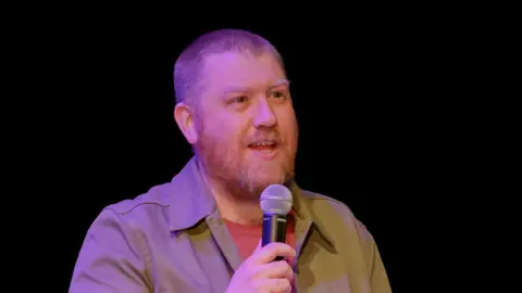 Sam Nicholls A man with short hair and beard talks into a microphone. He is wearing an orange T shirt and beige shirt