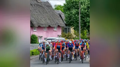 PA Media Ride London cyclists riding along a road, with the backdrop of a pink thatched cottage