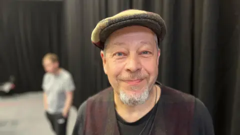 A man with a grey goatee and white hair, he is wearing a flat cap and a vest with a black t-shirt underneath