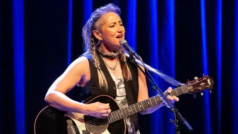 KT Tunstall performing on stage in front of a microphone and playing the guitar. She is wearing a black waistcoat and has her hair tied into plaits. 