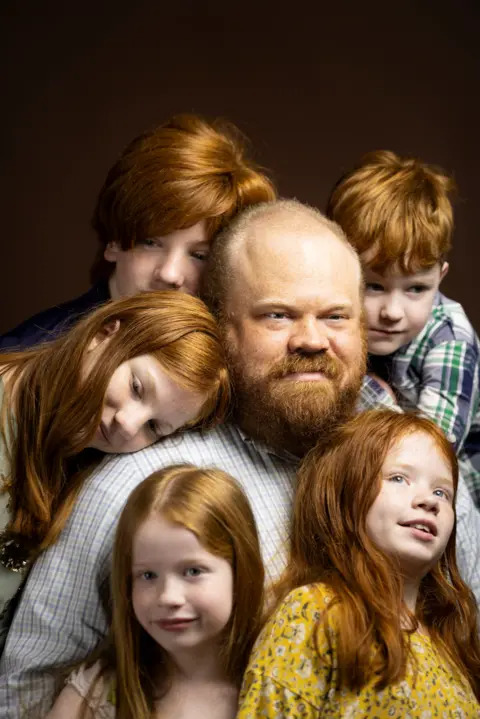Kieran Dodds Studio portrait of an adult with a beard seated in the center, surrounded by five children with red hair. Two children lean against the adult’s shoulders, one stands behind, and two are positioned in the foreground. The group is closely gathered against a dark brown background, with warm lighting highlighting their hair and patterned clothing.