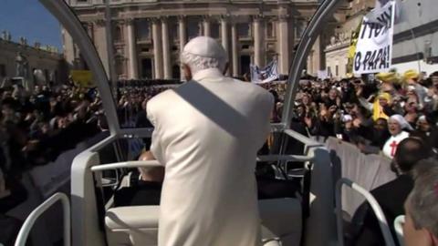 Pope Benedict XVI in the Popemobile facing away from the camera, surrounded by waving crowds