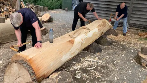 Operation Phoenix Three men, all wearing black T-shirts, chip away at a large log with tools