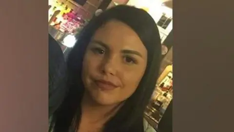 Alice Webb is smiling at the camera, she has dark hair and a piercing in her lower lip. She stands in front of an illuminated bar.