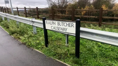 John Devine / BBC The side of a bridge with a silver railing and a black and white sign that reads "Ralph Butcher Causeway"