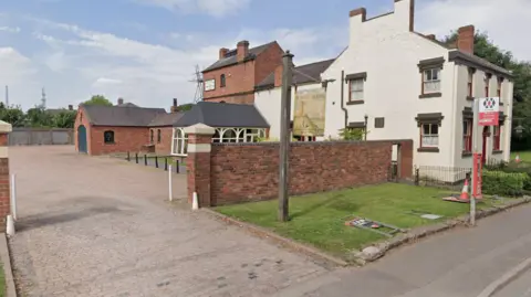 Google A cream painted building sits back from road and pavement, with a brick building and outbuildings behind it. There is a brick wall with an open entrance to a car park. There is a small metal fenced garden in front of the cream building, and to the side of it in front of the brick wall there is a sign for the Beacon Hotel and a bus stop. 