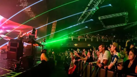 Getty Images Orang-orang di barisan depan konser, kanan, menonton Fatboy Slim di panggung di belakang set deck DJ, kiri